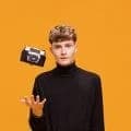 A young man in a black turtleneck tossing up a film camera against an orange wall