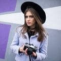 A woman in a black hat and lilac coat holding a camera in front of a purple wall