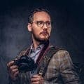 A bearded man in glasses with a camera over his shoulder against a dark background