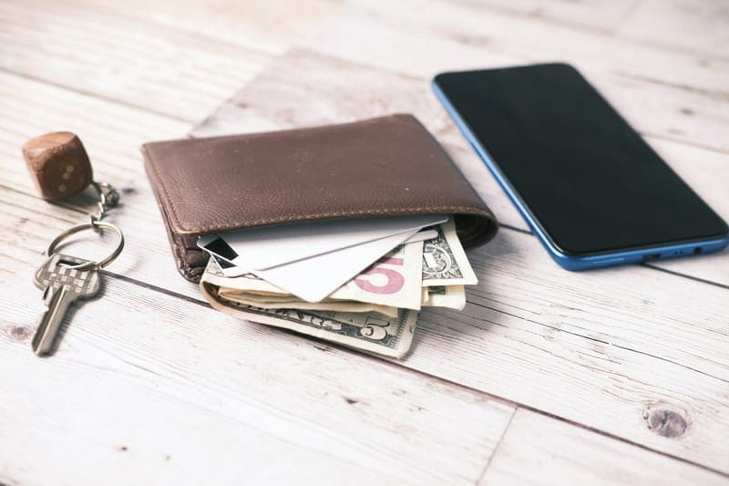 A leather wallet with dollar bills and cards beside a smartphone and keys