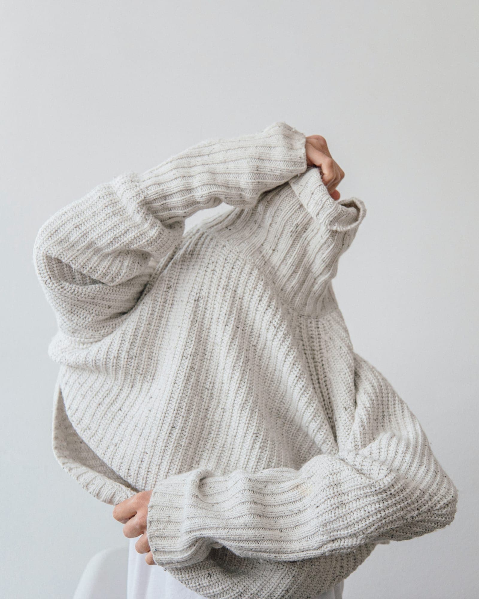 A person pulling a chunky cream knit sweater over their head
