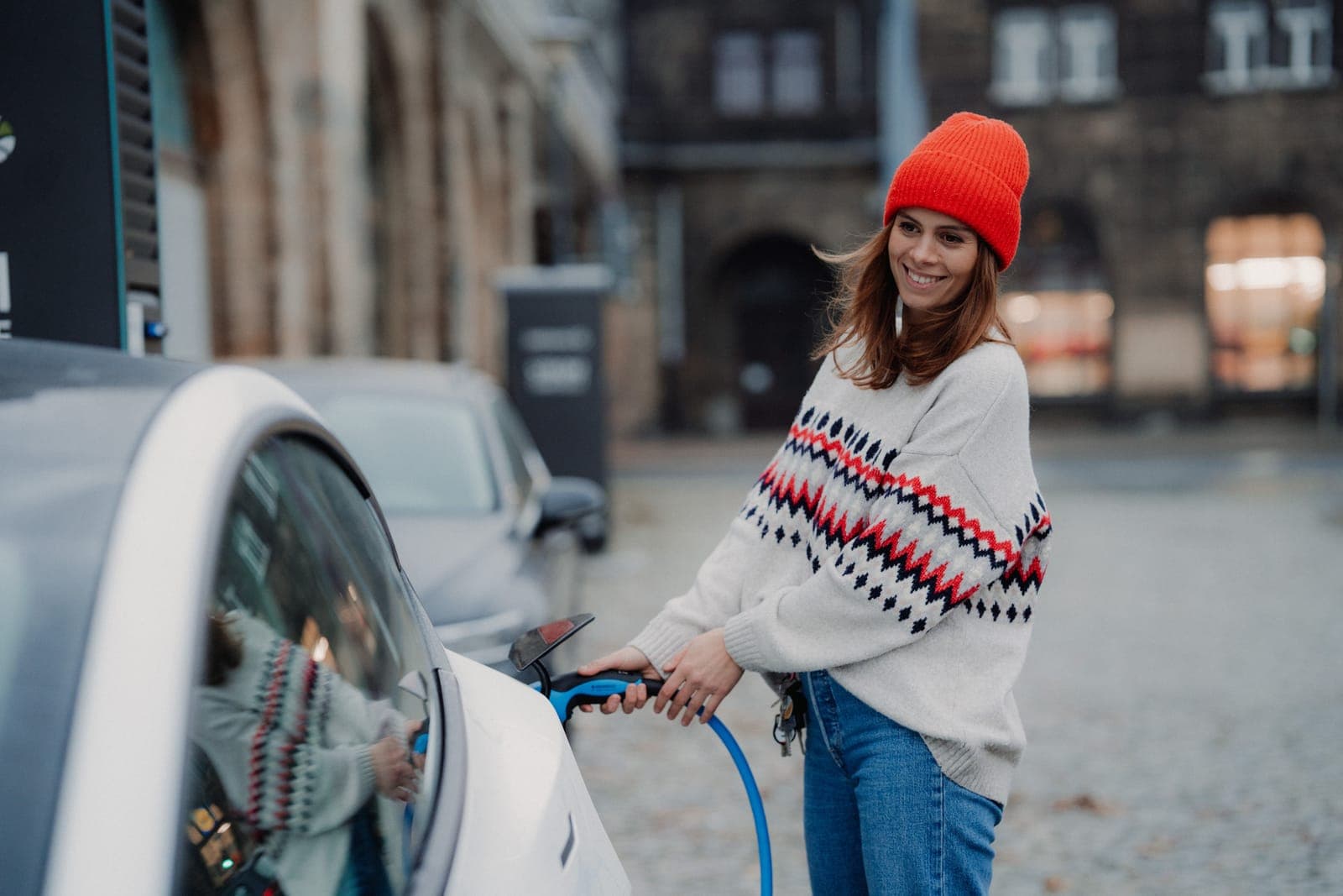 A smiling woman in a red beanie charging an electric car on a city street