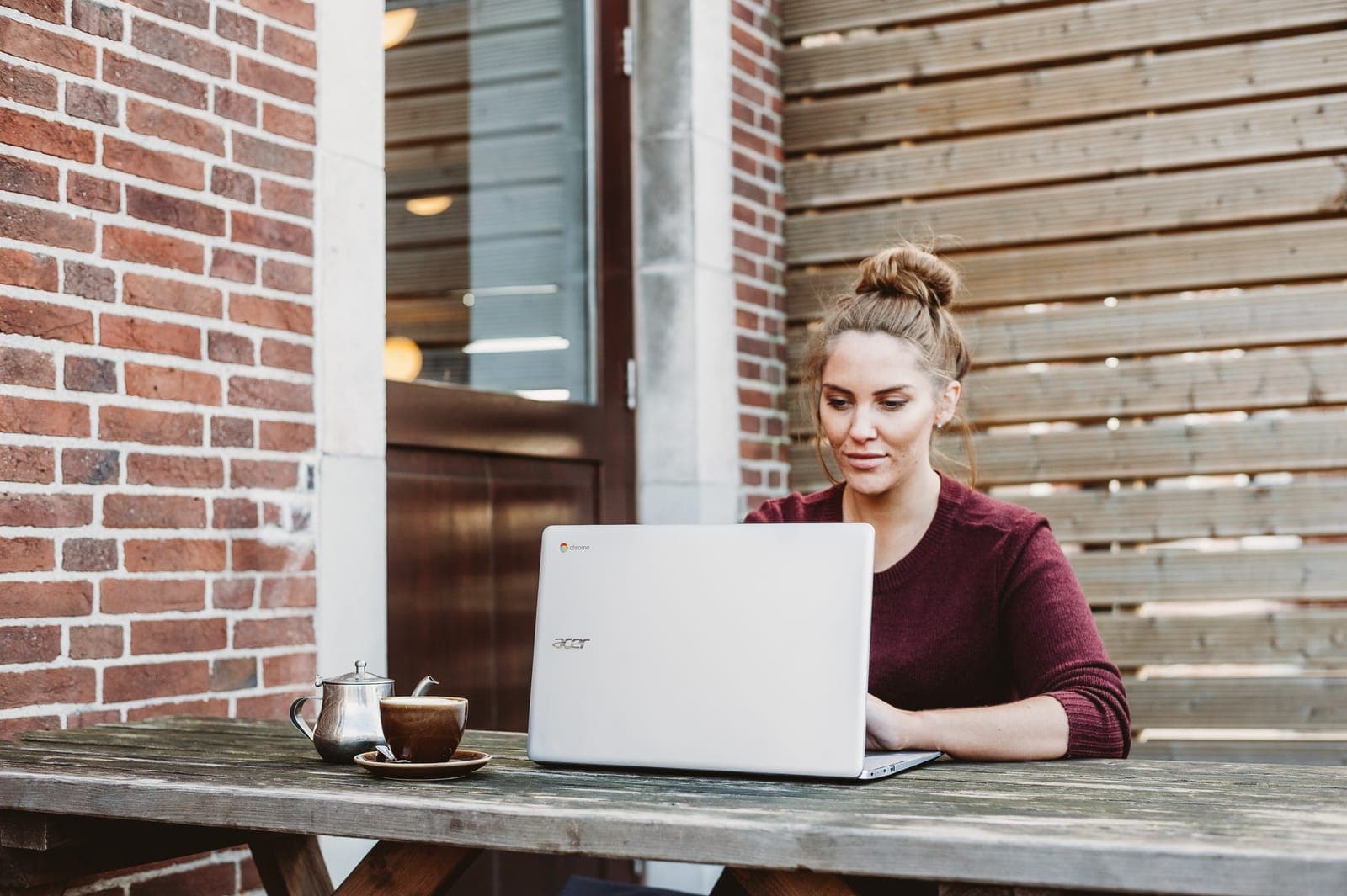 A woman working on a laptop at a wooden table outside a brick building
