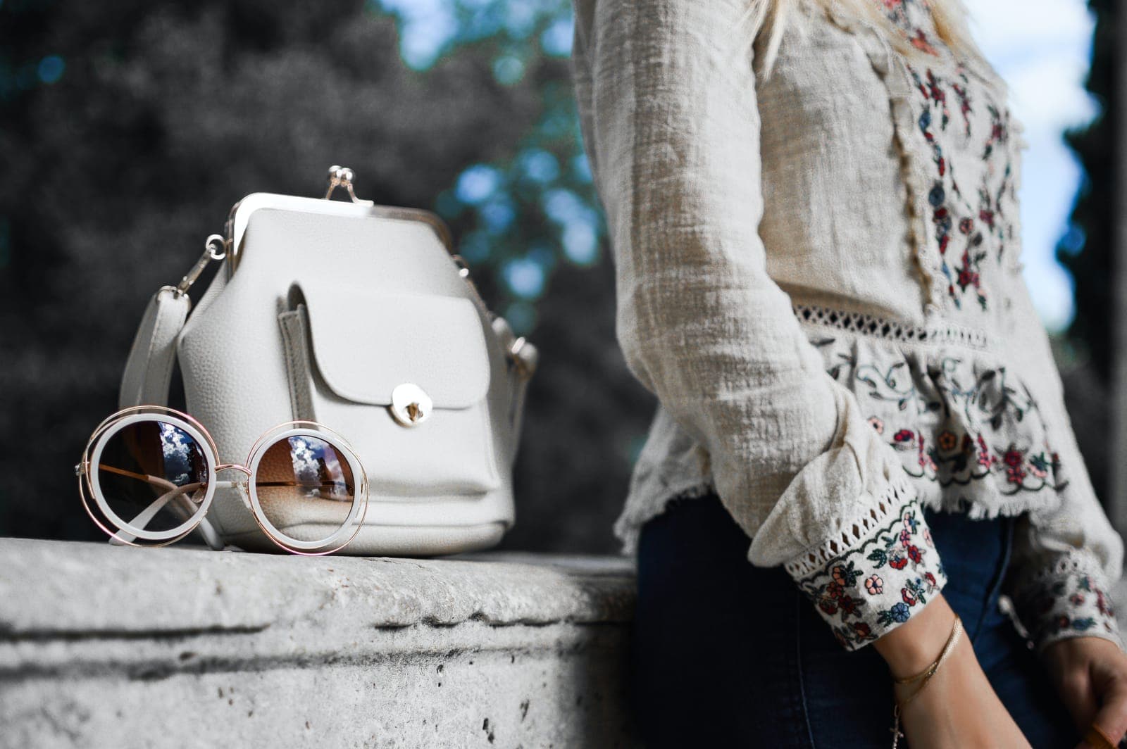 A woman in an embroidered blouse beside a white handbag and round sunglasses